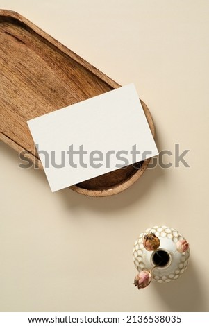 Blank paper card mockup on wooden tray with vase of dried flowers on beige background. Flat lay, top view, copy space.