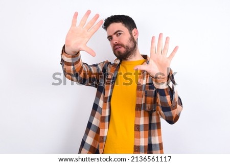 Portrait of smiling young caucasian man wearing plaid shirt over white background looking at camera and gesturing finger frame. Creativity and photography concept.