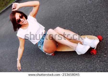Pretty young smiling happy laughing girl dressed in jeans shorts and white T-shirt sitting on long-board and having fun. Downhill, longboarding . Outdoors, lifestyle. 