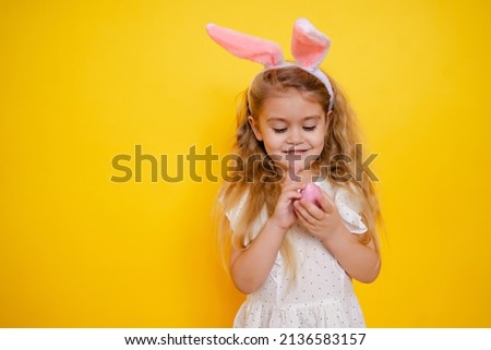 cute smiling blonde girl with bunny ears holding an easter egg in her hands, on a yellow background studio, space for text, kid celebrate easter