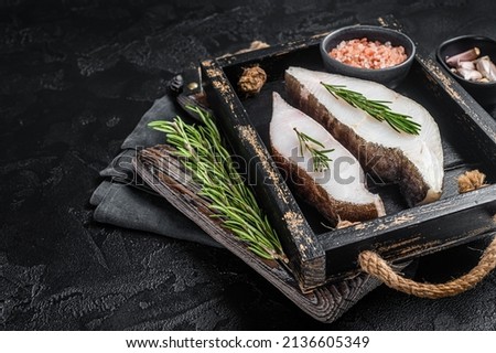 Raw halibut fish steak in wooden tray with herbs. Black background. Top view. Copy space
