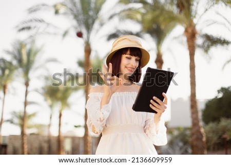 Happy traveling woman on white dress and straw hat using tablet reading news on the Internet using wifi in exotic hotel. Woman working on the digital tablet showing sign ok