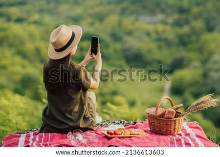 Pretty young woman in hat and dress is using smart phone, take picture while sitting at outdoor picnic. 