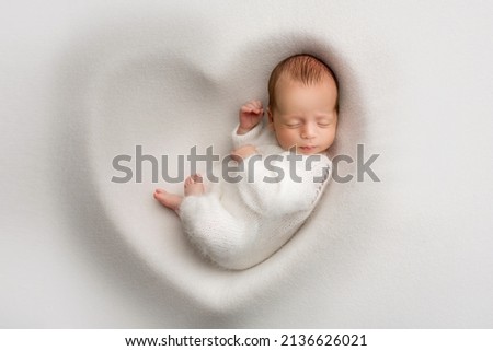 A sleeping newborn boy in the first days of life in a white soft cocoon with a knitted woolen white hat on a white background in the shape of a heart. Studio macro photography, portrait of a newborn.