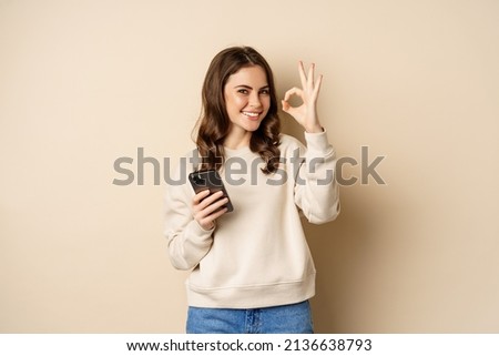 Beautiful woman holding cellphone, mobile phone and okay sign, recommending application, shopping app, standing over beige background