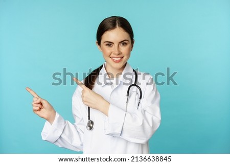 Portrait of smiling medical worker, girl doctor in white coat with stethoscope, pointing fingers left, showing medical clinic advertisement, torquoise background