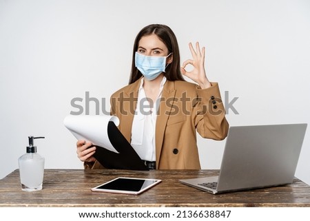 Woman working in office at table, holding clipboard with papers, showing okay sign, wearing medical face mask, white background