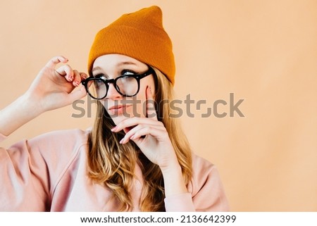 Portrait of a blonde hair girl with wearing glasses and an orange hat. The girl is posing against the light orange background