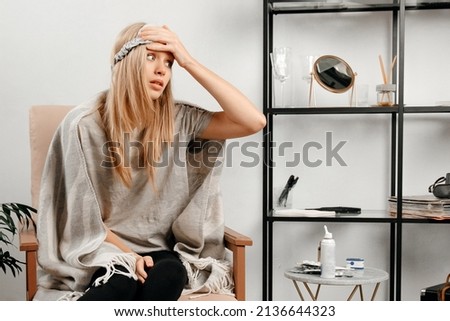 Blonde hair girl with a sleeping mask on the face is sitting in a chair. Young woman is holding a hand on the head because she has strong headache