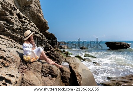Girl in a white dress and a straw hat sits on the rocks and looks at the sea. Healing meditation. Summer heat. Holidays.
