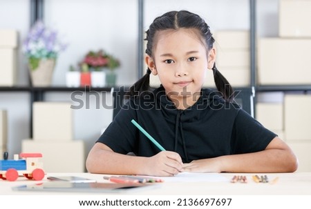 Asian young smart clever elementary schoolgirl artist in casual outfit sitting using color pencils drawing painting picture on paper alone on working table with tablet and wooden toy in living room.