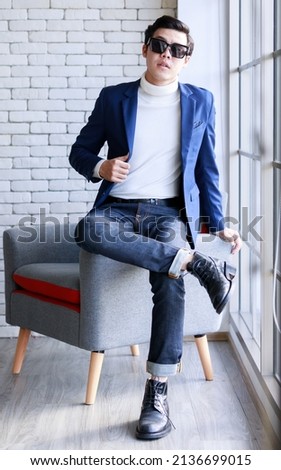 Studio shot Asian young smart handsome successful professional male businessman in casual blue suit turtle neck shirt sunglasses sit on armchair looking at camera on brick wall background Royalty-Free Stock Photo #2136699015