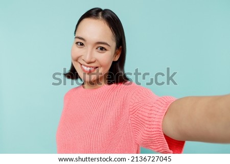 Close up young happy woman of Asian ethnicity 20s in pink sweater doing selfie shot pov on mobile phone isolated on pastel plain light blue color background studio portrait. People lifestyle concept