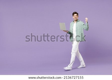 Full length body young fun happy smiling man in casual mint shirt white t-shirt use laptop pc computer do winner gesture clench fist walk isolated on purple background studio. People lifestyle concept