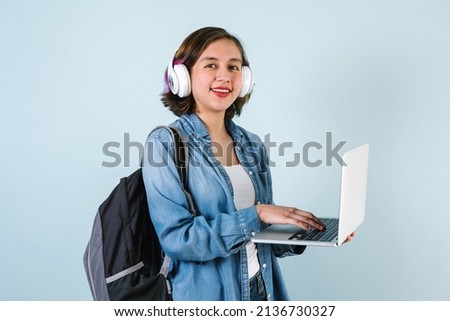 Young Hispanic student woman listen music with headphones and holding computer over isolated blue background