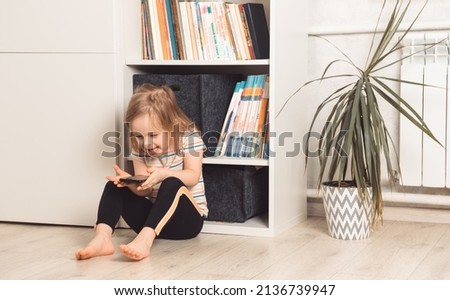 Little child sits on the floor in his room and uses the phone. Smiling girl looking at the phone. Childhood communication, technology and communication concept
