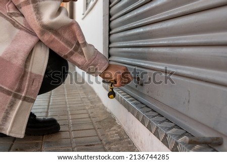 A close up shot  of a young woman lowering the metal fence of her business at the time of closure. Brunette girl crouching to close her sales room once her working day is over Royalty-Free Stock Photo #2136744285