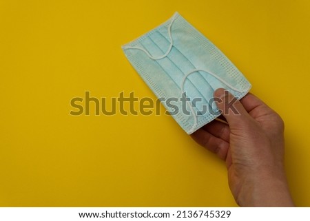Close-up image of a hand holding a blue face mask on a yellow background leaving copy space. Sanitary hygiene concept in times of viruses and diseases.