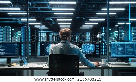 Software Developer Works with AI Analysis on his Computer with Two Monitors. Developer Coding while Looking at the Display of Computer. Modern Evening Dark Office. Back view. Royalty-Free Stock Photo #2136788215