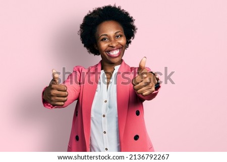 African american woman with afro hair wearing business jacket approving doing positive gesture with hand, thumbs up smiling and happy for success. winner gesture.  Royalty-Free Stock Photo #2136792267
