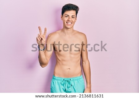 Young hispanic man wearing swimwear shirtless showing and pointing up with fingers number two while smiling confident and happy. 