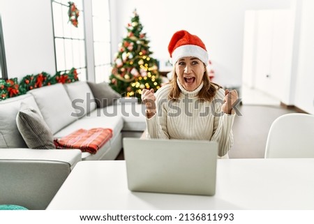 Middle age woman wearing santa claus hat using laptop celebrating mad and crazy for success with arms raised and closed eyes screaming excited. winner concept 