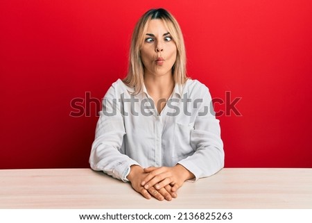 Young caucasian woman wearing casual clothes sitting on the table making fish face with lips, crazy and comical gesture. funny expression. 