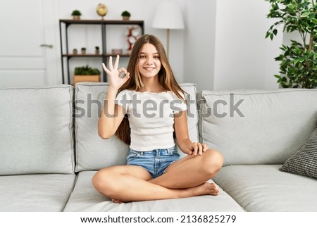 Young brunette teenager sitting on the sofa at home smiling positive doing ok sign with hand and fingers. successful expression. 