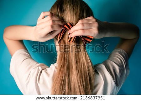 St. George Ribbon. The girl ties a St. George ribbon on her hair. Blonde girl. On a blue background. Victory. Tying up hair.Portrait photograph of a young girl.