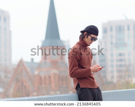 Portrait of handsome Chinese young man with sunglasses standing and typing at mobile phone with Shanghai city landmarks background, male fashion, cool Asian young man lifestyle.
