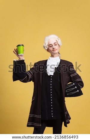 Coffee. Portrait of young elegant man in white wig and vintage medieval outfit posing isolated on yellow background. Art, beauty, fashion. Retro style, eras comparison. Copy space for ad