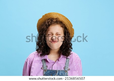 Emotional young curly girl wearing plaid shirt and hat making faces isolated on blue background. Charming female model in image of gardener. Concept of funny meme emotions, facial expressions