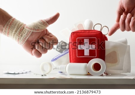 Hand pointing at travel portable first aid bag full of medical objects and tools with bandaged hand with ok sign. Front view. Horizontal composition