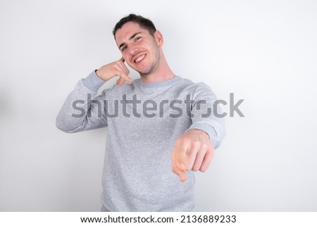 young handsome caucasian man wearing grey sweater over white background smiling cheerfully and pointing to camera while making a call you later gesture, talking on phone