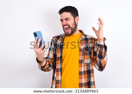 Photo of outraged annoyed young caucasian man wearing plaid shirt over white background holds cell phone, makes call, argues with colleague,  expresses negative emotions. People and anger. Royalty-Free Stock Photo #2136889865