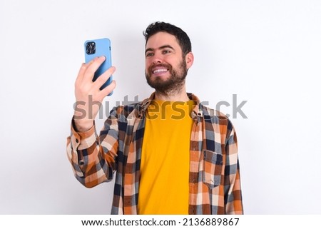 Isolated shot of pleased cheerful young caucasian man wearing plaid shirt over white background, makes selfie with mobile phone. People, technology and leisure concept