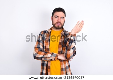 Studio shot of frustrated young caucasian man wearing plaid shirt over white background gesturing with raised palm, frowning, being displeased and confused with dumb question.
