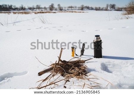 The thermos stands on the snow against the background of the forest. A place to light a fire, prepare for cooking in the wild. High quality photo