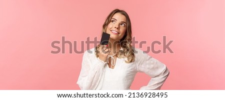 Close-up portrait of thoughtful and creative blond woman in white dress, touching chin with credit card and smiling dreamy as looking up and thinking, picturing what buy as present, pink background