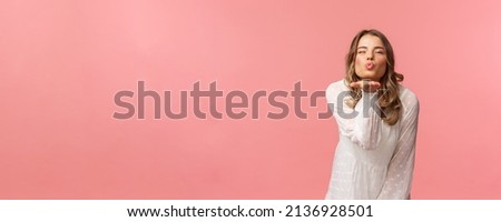 Beauty, fashion and women concept. Portrait of romantic tender, young blond woman in white trendy dress, sending air kiss at camera with sassy flirty smile, hold hand near lips, pink background