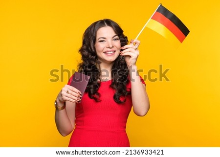 Young woman in red dress showing small flag Germany and passport on yellow backdrop in studio. Study abroad concept. International student exchange program. Learning German Deutsch with native speaker Royalty-Free Stock Photo #2136933421