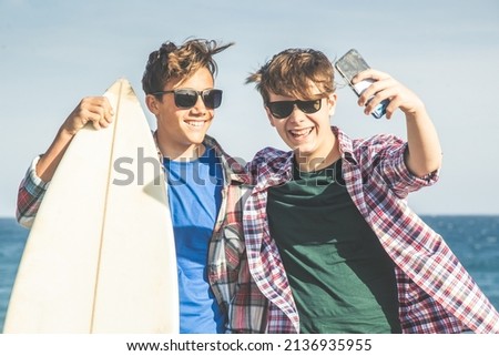 Two young boys on the beach near ocean taking picture with cell phone, schoolmates on vacation in the easter break amuse themselves on the coast with surf. Video chat with friends wifi anywhere.