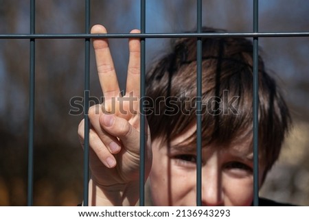 Child refugee showing peace sign behind a metal fence. Social problem of war migrants.