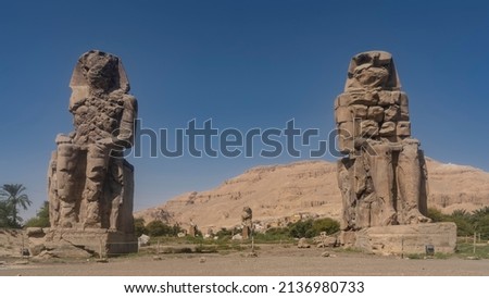 Giant stone sculptures of the Colossi of Memnon in Egypt. Huge statues of seated pharaohs against a clear blue sky and sand dunes.