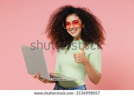 Fun young curly latin woman 20s years old wears mint t-shirt sunglasses hold use work on laptop pc computer showing thumb up like gesture isolated on plain pastel light pink background studio portrait