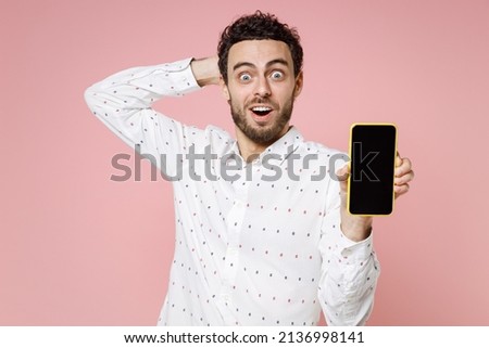 Shocked amazed young bearded man 20s wearing basic casual white shirt standing hold mobile cell phone with blank empty screen put hand on head isolated on pastel pink color background studio portrait