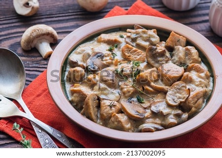 Stewed diced turkey with mushrooms in a creamy sauce in a clay plate on a brown wooden background. Turkey recipes. Second hot dishes