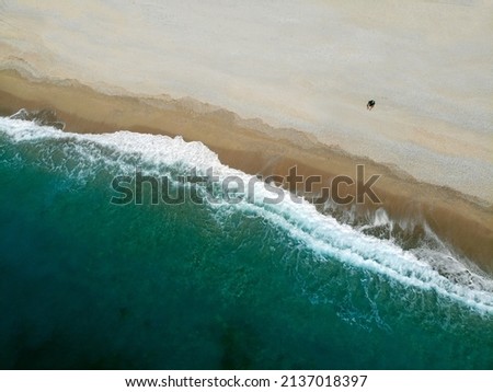 Drone aerial picture view of one man sitting on fine sand with little white waves and blue green turquoise water with corals of Gipje beach. Top down view of one person on beach.