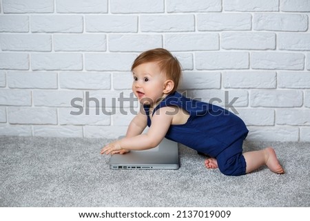 A small child 6-7 months old in a blue cotton overalls sits with an open laptop and watches educational games