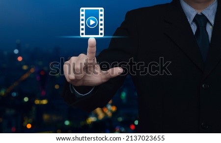 Businessman pressing play button with movie flat icon over blur colorful night light modern city tower and skyscraper, Business cinema online concept
